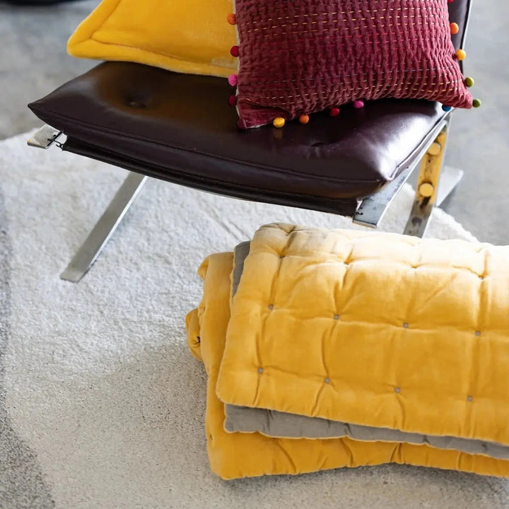 Chemin de lit Vivaraise jaune matelassé plié au sol près d'un fauteuil en cuir noir avec coussins colorés
