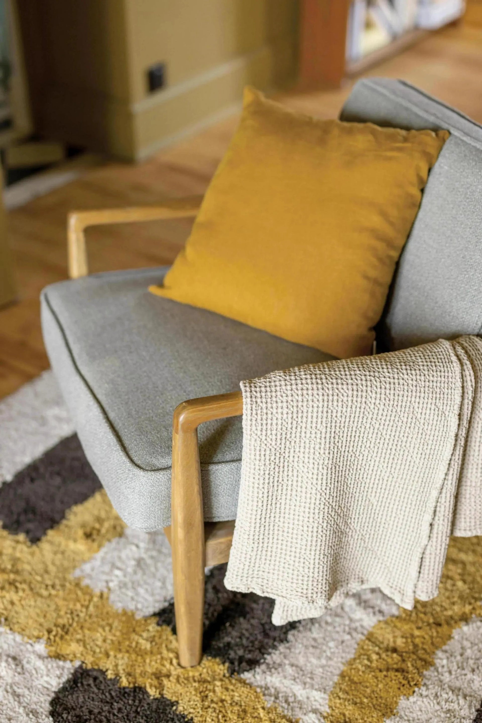 Plaid beige en maille douce Vivaraise posé sur un fauteuil gris avec un coussin jaune