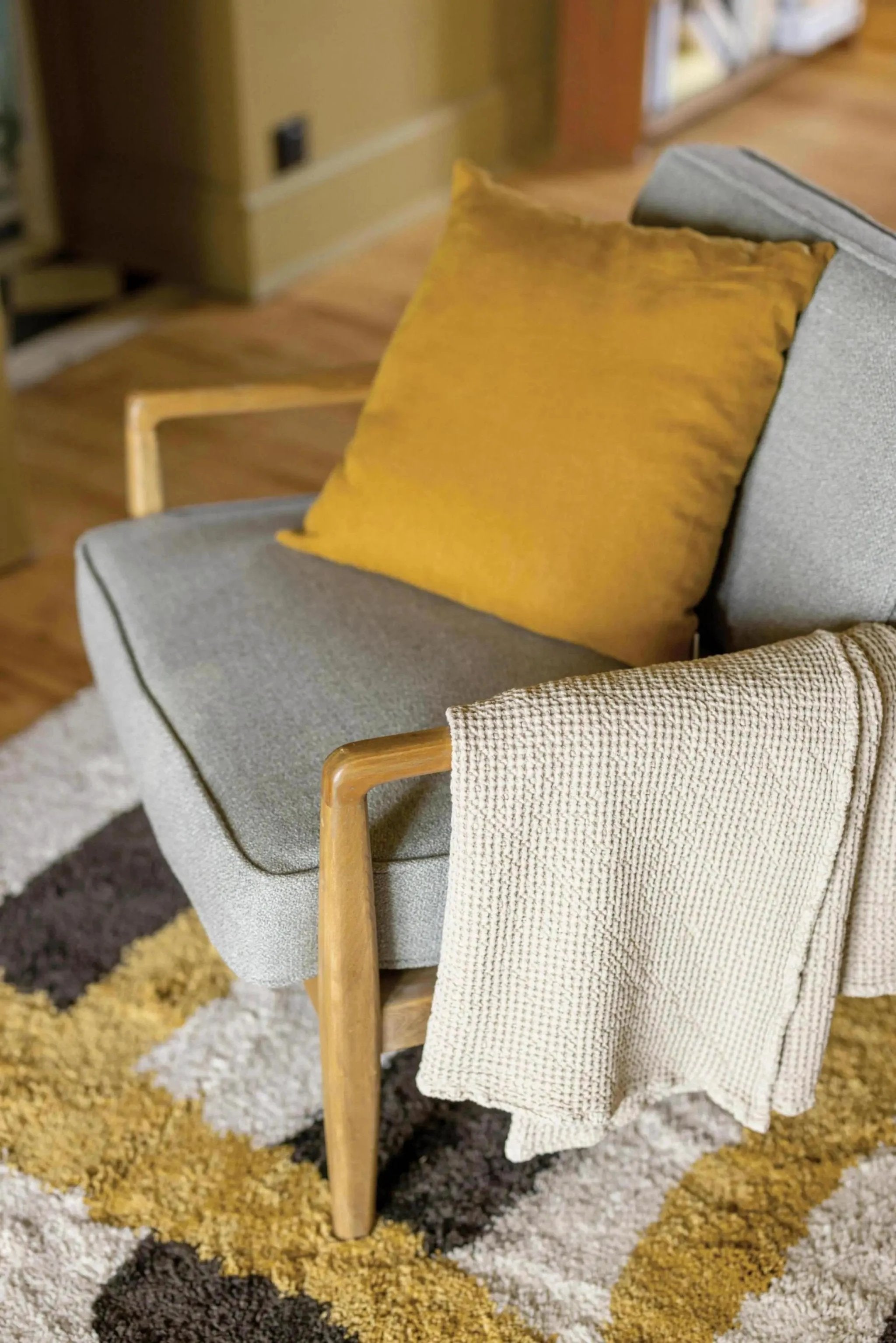 Plaid beige en maille douce Vivaraise posé sur un fauteuil gris avec un coussin jaune