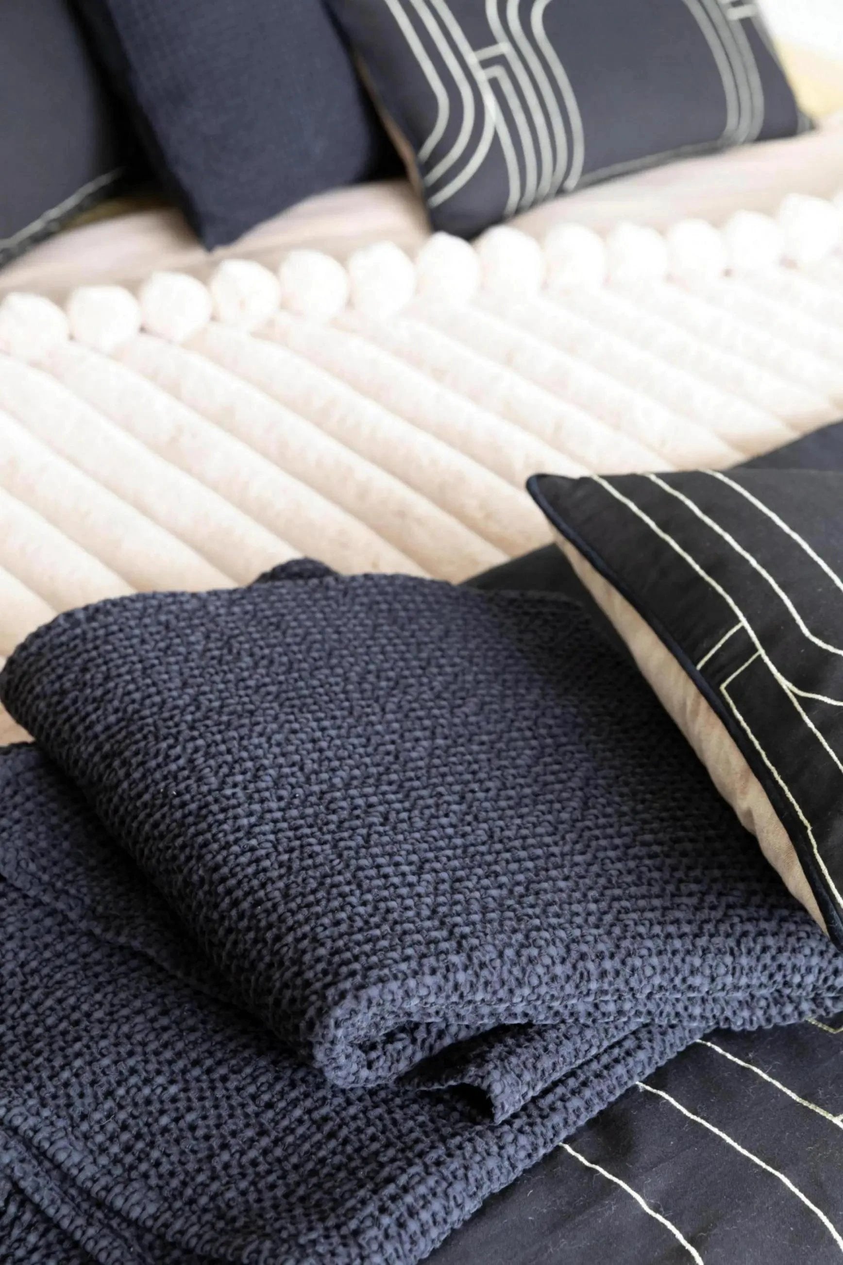 Plaid Vivaraise bleu marine en laine texturée plié sur un lit avec coussins noirs et crème
