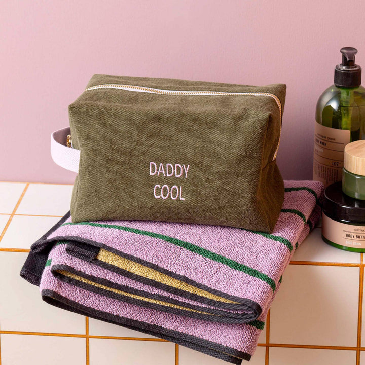 Trousse kaki "Daddy Cool Kale" posée sur des serviettes violet à rayures, près de produits de soin.