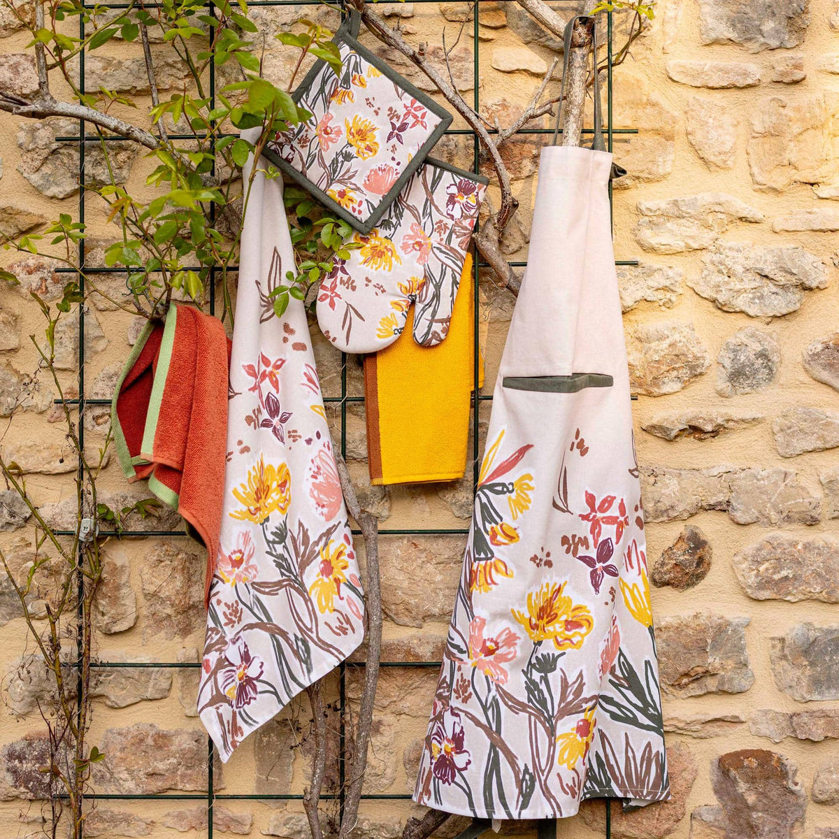Torchon et gants en coton imprimé floral coloré suspendus devant un mur en pierre, avec torchon et tablier assortis. Torchon Skica coton imprimé.