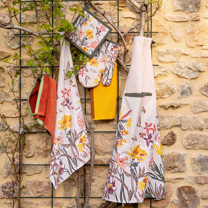 Torchon et gants en coton imprimé floral coloré suspendus devant un mur en pierre, avec torchon et tablier assortis. Torchon Skica coton imprimé.